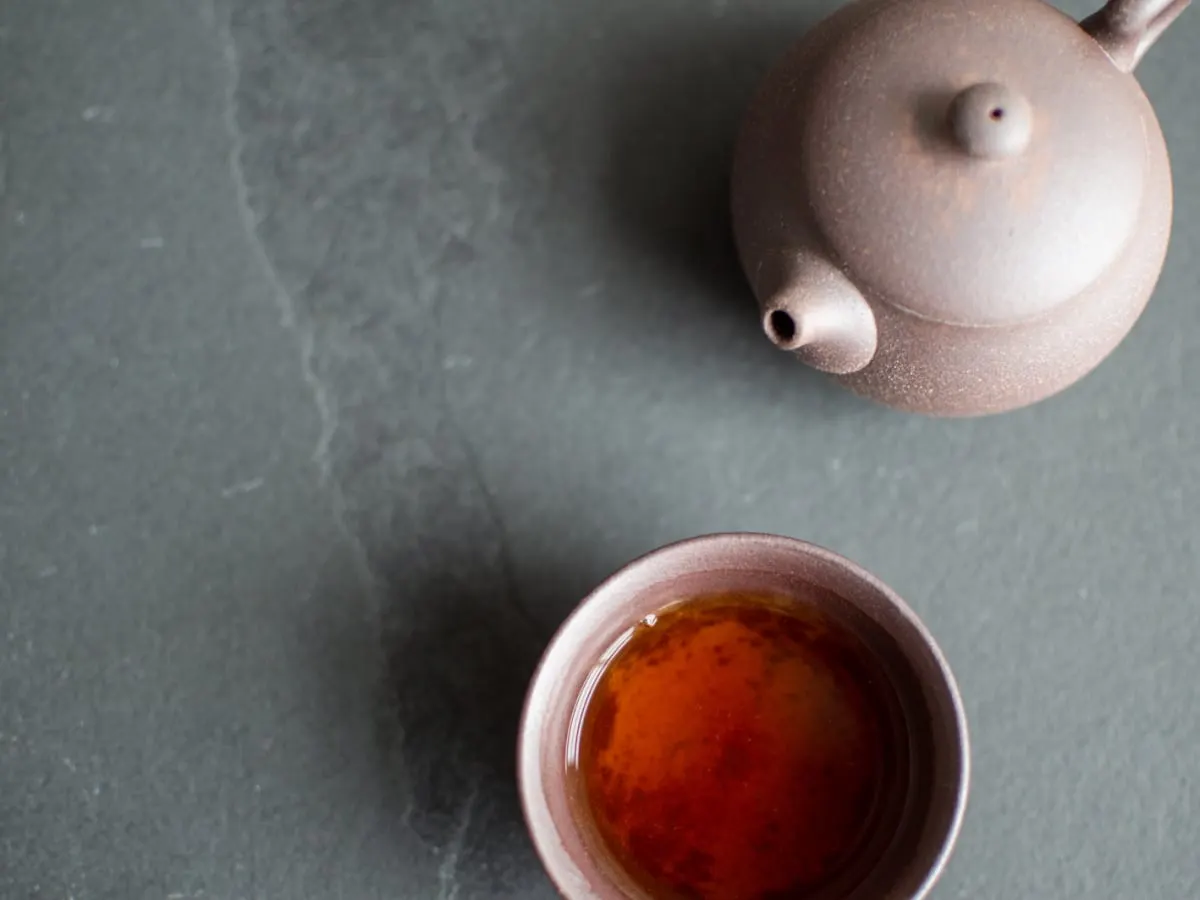 Une théière en argile et une tasse remplie de thé ambré reposent sur une surface sombre et épurée. L’image évoque un moment de calme, de découverte et de partage autour de l’univers du thé.