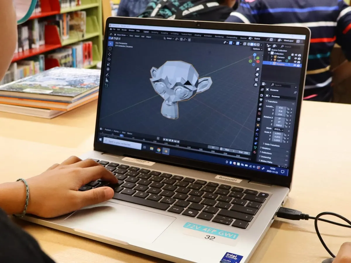 Gros plan sur un ordinateur : on voit le modèle 3D d'un visage de singe à travers le logiciel de modélisation 3D Blender. On voit des mains manipuler une souris et le clavier de l'ordinateur.
