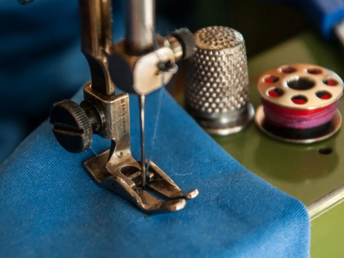 Pied de biche de machine à coudre déposé sur tissu bleu, dé et roulette de fil déposés à côté.