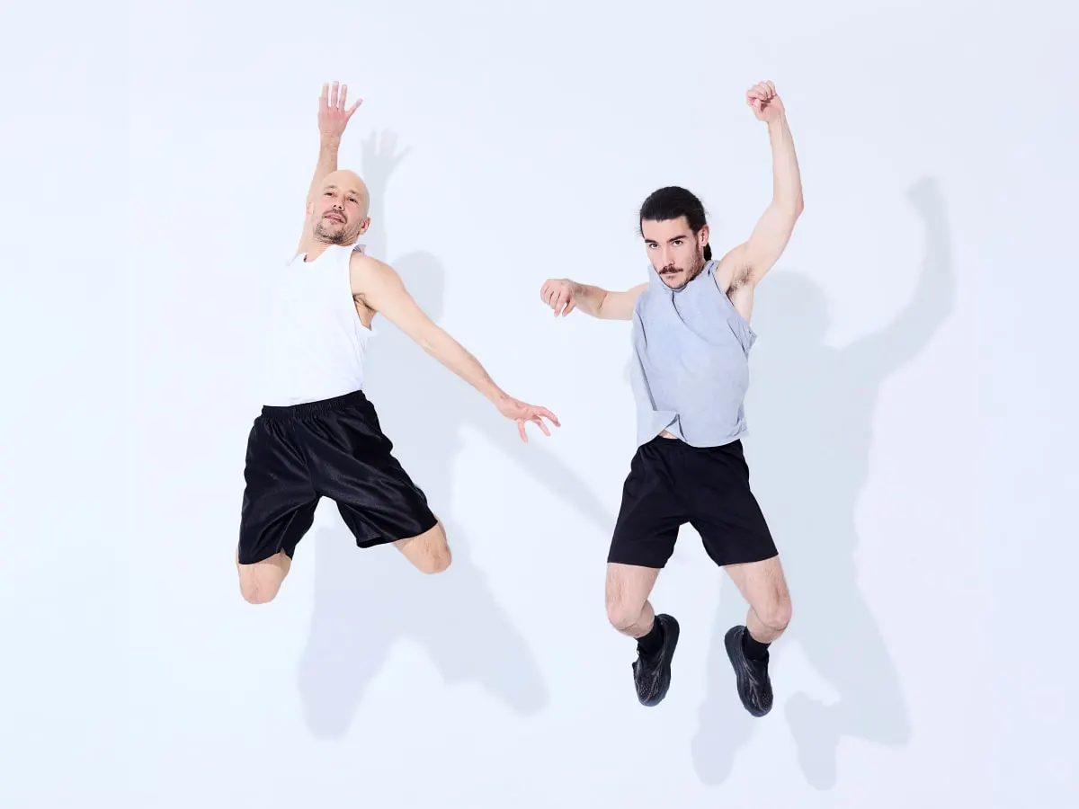 Deux danseurs en mouvement sautent sur fond neutre dans une composition minimaliste et dynamique.