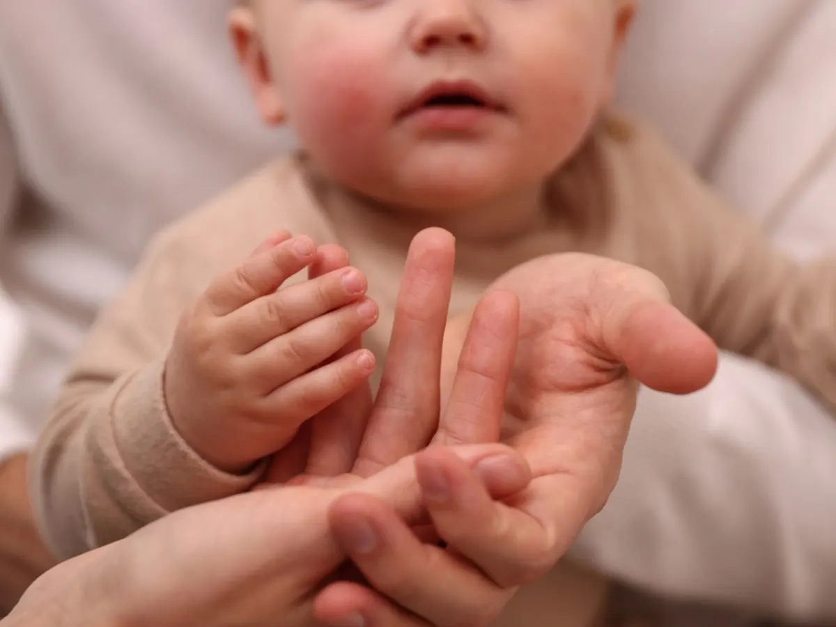 Gros plan sur les mains d’un bébé qui touche et explore les doigts d’un adulte, tandis qu’une petite maraca en bois est visible dans une autre main. L’image évoque la communication précoce, l’éveil sensoriel et l’interaction entre le bébé et son entourage.