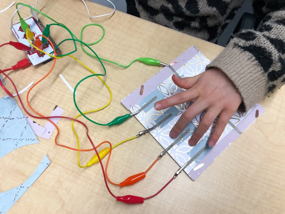 On voit la main d'un enfant manipuler un bricolage techno. Des pinces crocodiles de toutes les couleurs sont branchées à un petit piano fait de papier d'alluminium. Il s'agit du microcontrôleur Makey Makey.