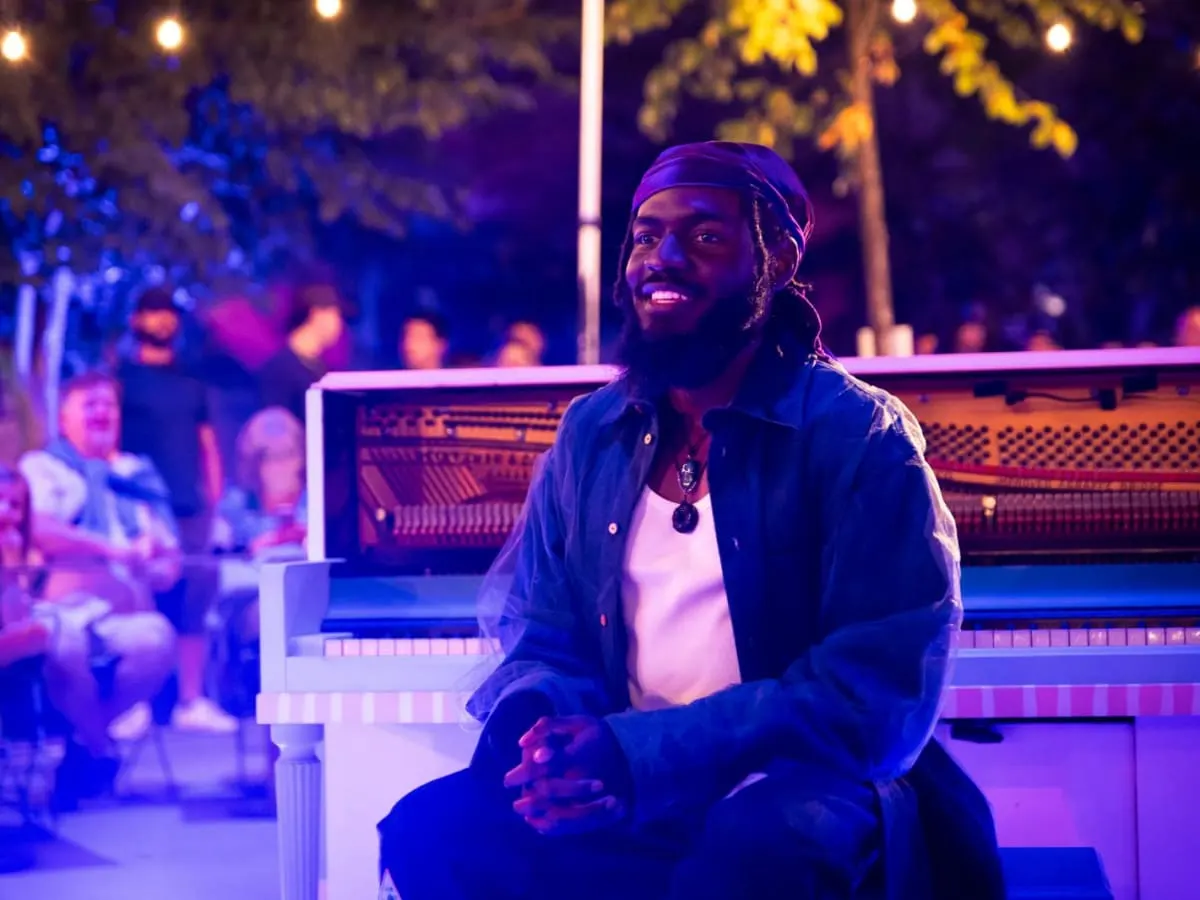 La photo présente l'artiste souriant devant son piano dans un éclairage violet
