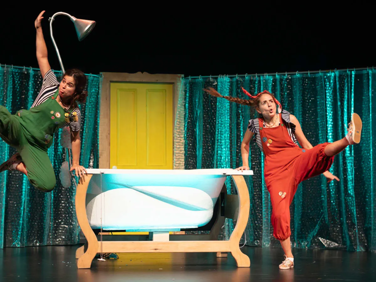 Deux comédiennes en salopettes colorées sautent joyeusement de chaque côté d'une baignoire sur une scène de théâtre pour enfants.