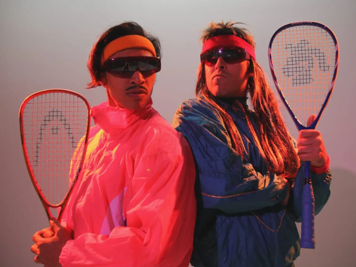 Deux hommes portant des habits des années 80 et munis d'une raquette de tennis