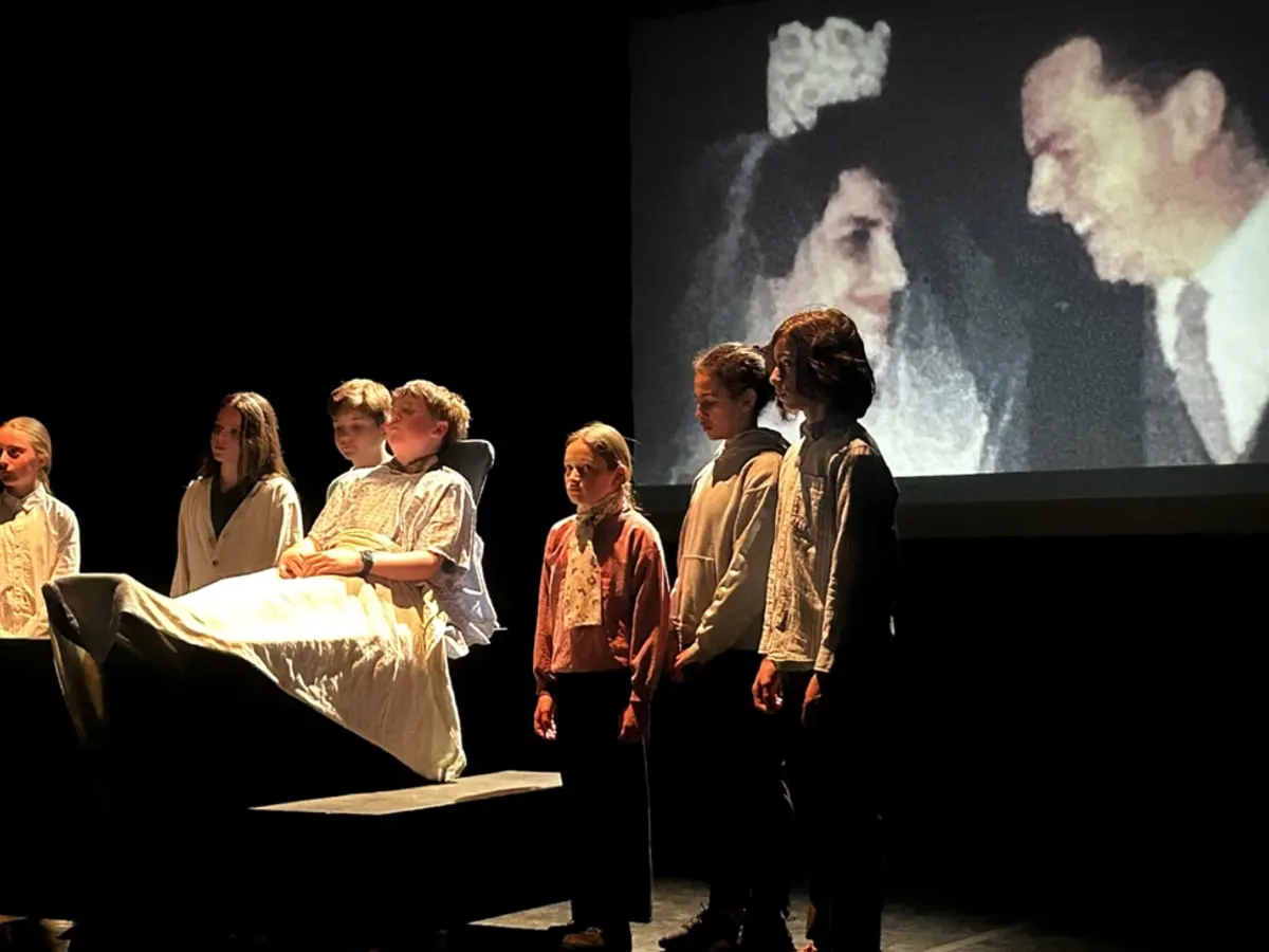 Sur une scène de théâtre, plusieurs enfants entourent un lit devant une grande projection d’une photographie.