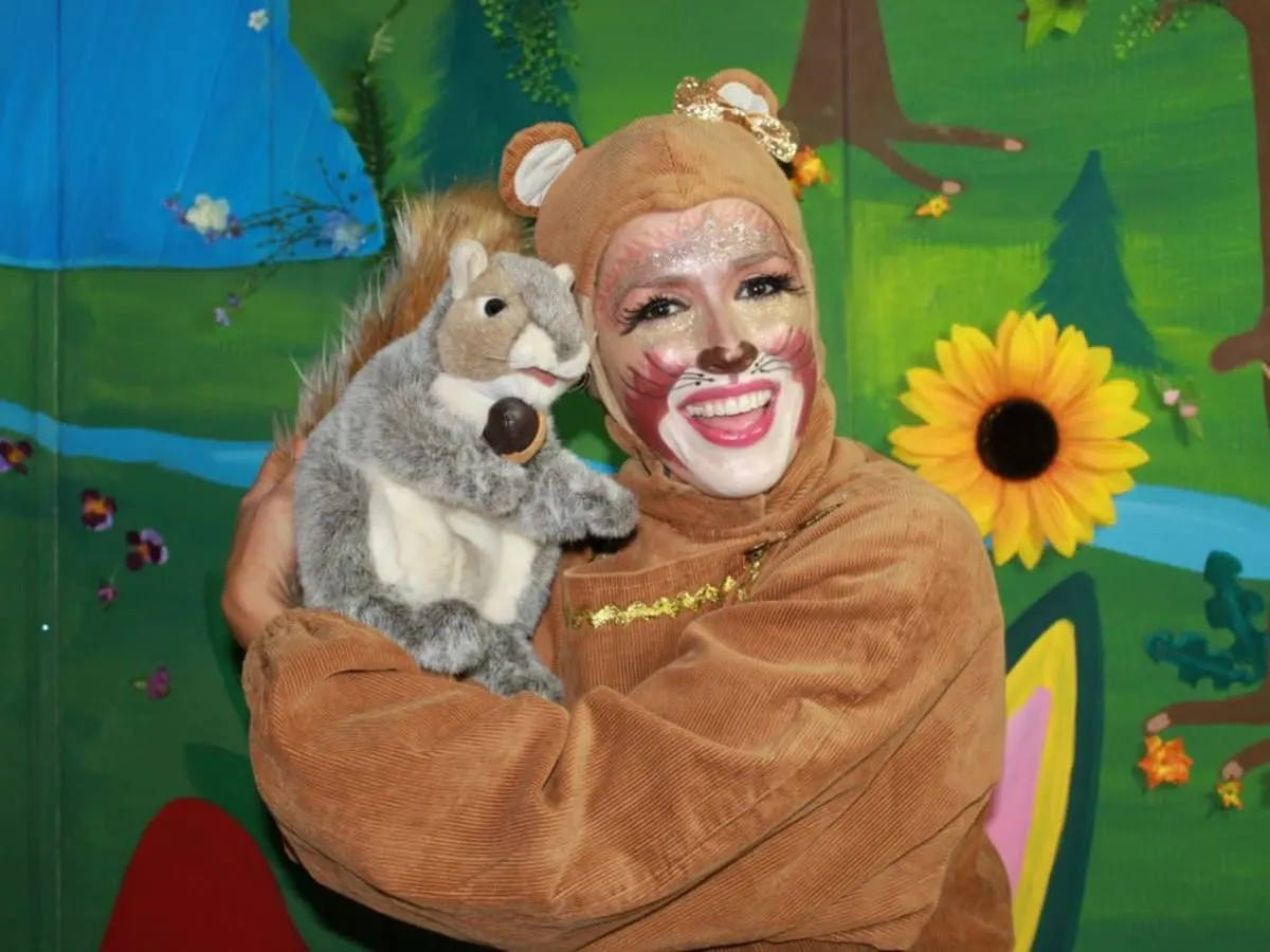 Une personne costumée en petit animal des bois, avec le visage maquillé et une tenue brun clair, tient une marionnette d’écureuil. Le décor coloré représente une forêt imaginaire avec des arbres, des fleurs et un ruisseau, créant une ambiance festive et familiale.