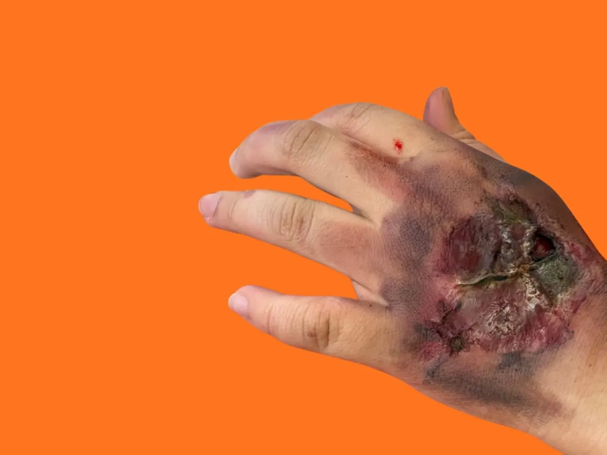 Gros plan d'une main avec une fausse blessure devant un fond orange vif. L’image met en valeur une création d’effets spéciaux originale et ludique inspirée de l’univers du maquillage et des accessoires fantastiques..