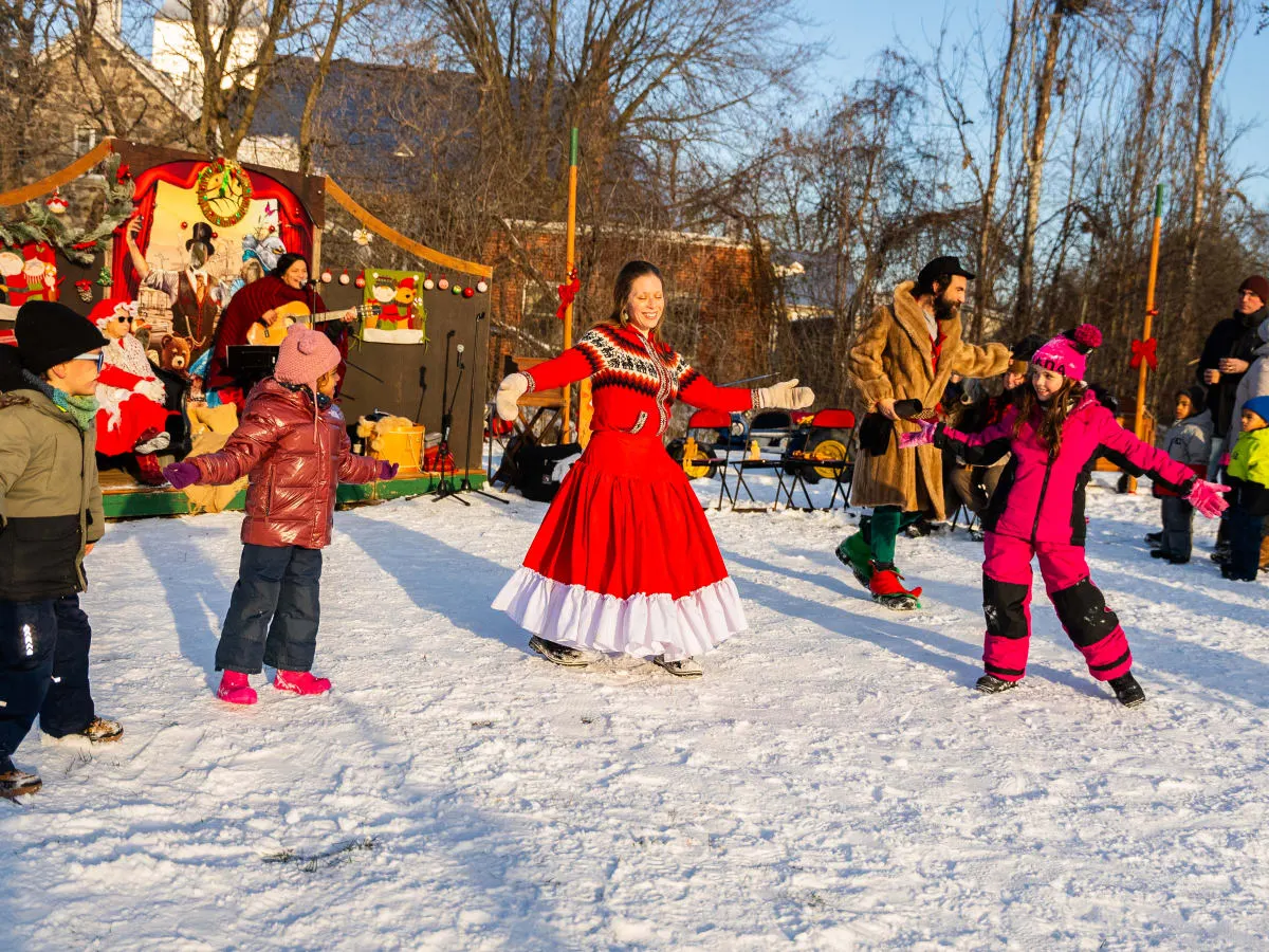 Enfants participant à une animation extérieure de Noël dans un parc enneigé, devant une scène festive avec musique et décorations du temps des Fêtes.