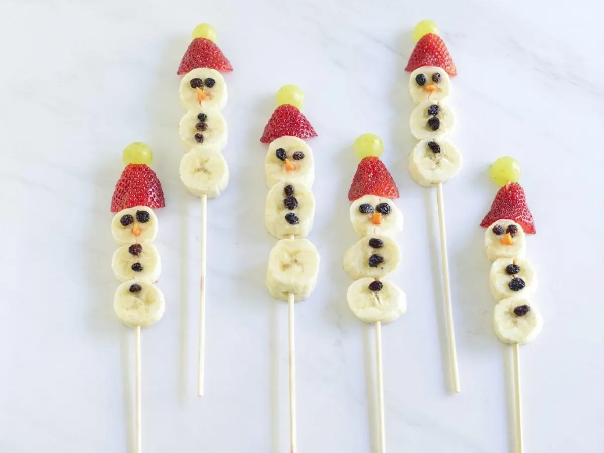 Cette image montre cinq brochettes de fruits décorées en bonshommes de neige, disposées verticalement sur une surface blanche à l'aspect marbré.