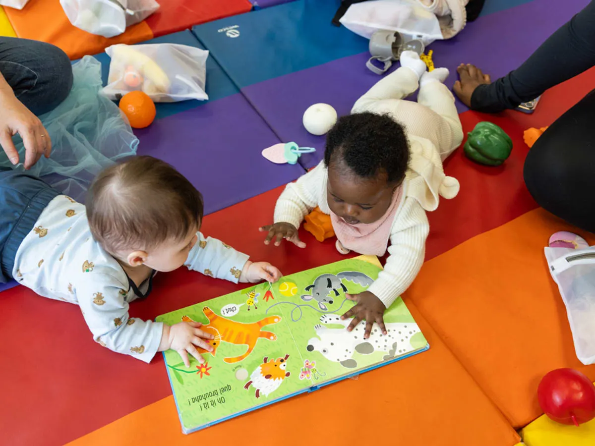 Deux bébés à plat ventre, sur un tapis de sol coloré, qui regardent un livre et qui sont entourés de jouets sensoriels.